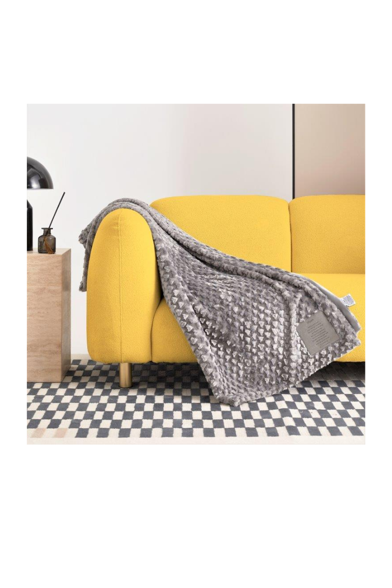 A grey blanket, with a patch that can be personalized, draped over the arm of a yellow couch. 