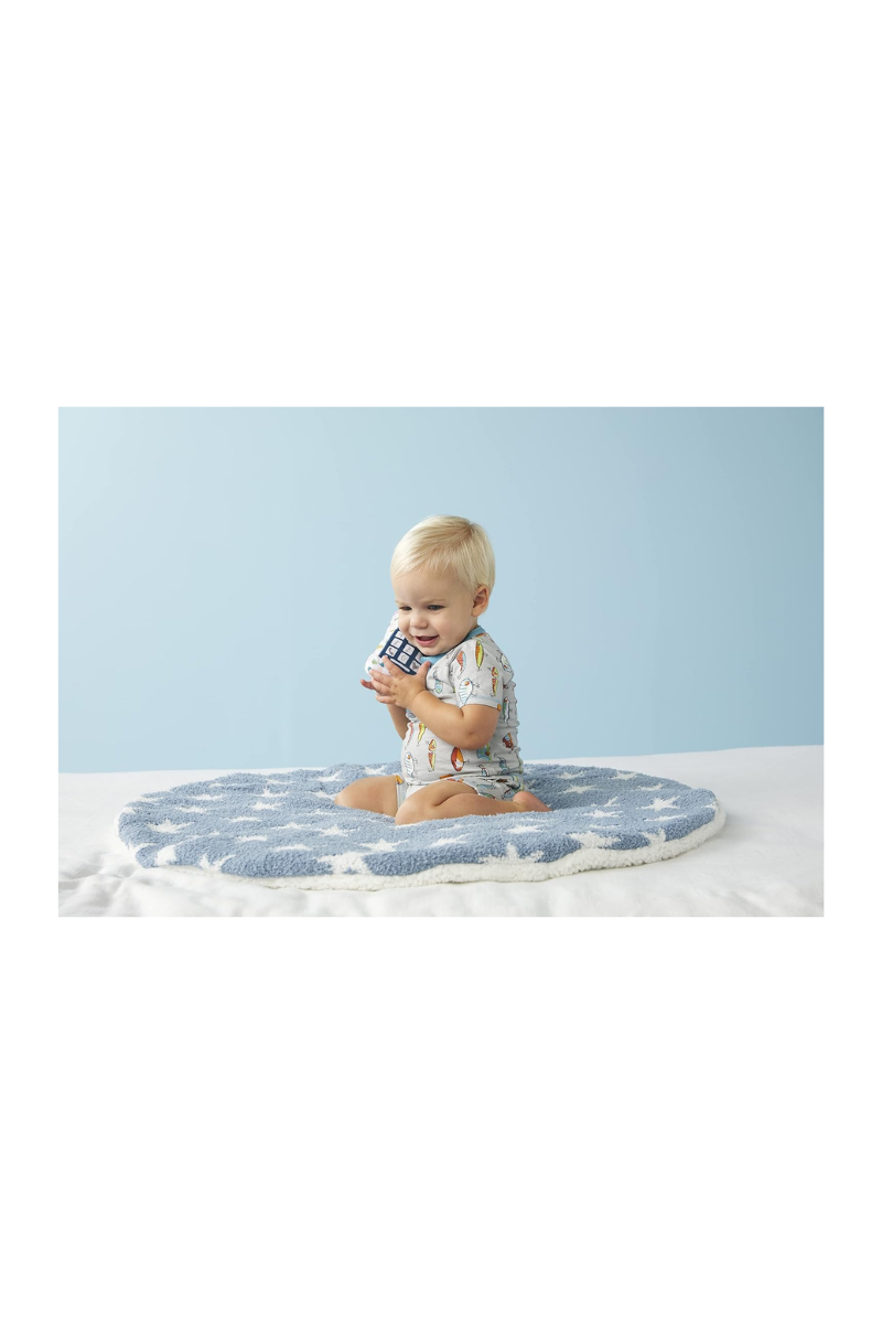 A little boy with blonde hair sitting on a blue circle mat with stars on it. He is playing with a plush phone. The floor is white and the wall behind him is blue.