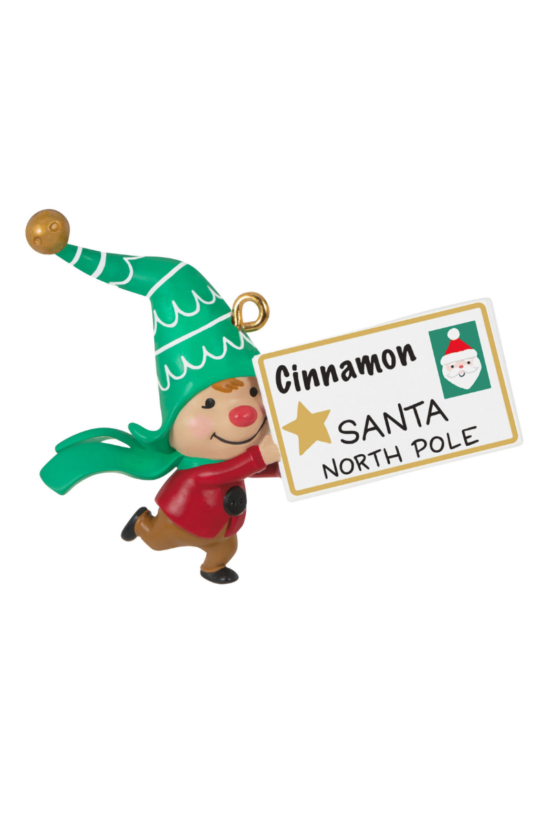 A Christmas ornament depicting an elf wearing a red coat and a green hat and scarf running holding a letter addressed to Santa from "Cinnamon".
